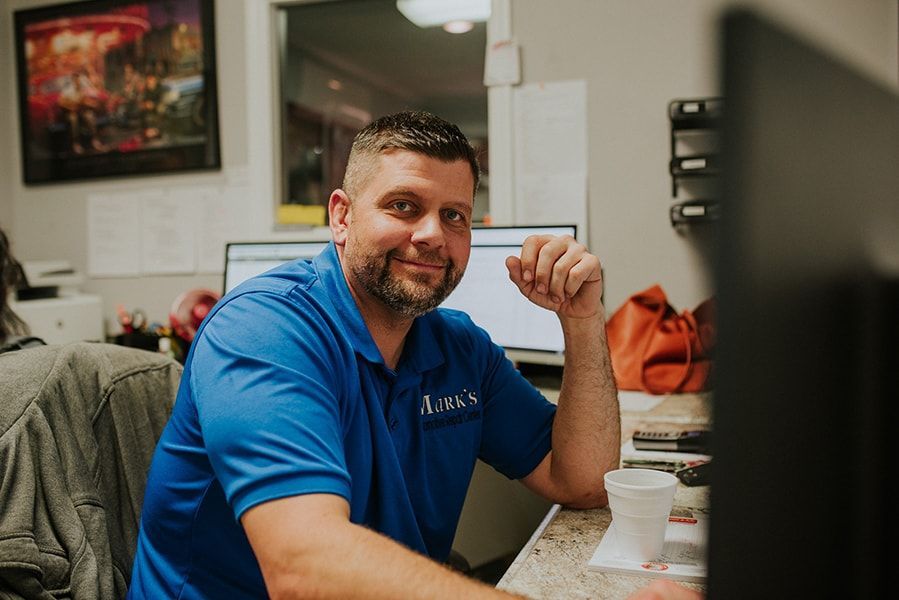 Man in blue shirt, smiling at desk with computer and coffee cup. | Emge's Car Craft Auto Services