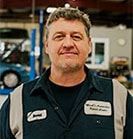 Man in mechanic uniform smiling in a garage; a car is in the background. | Emge's Car Craft Auto Services