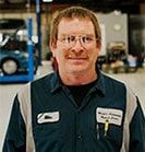 Mechanic in blue and gray work uniform smiles in a garage. | Emge's Car Craft Auto Services
