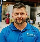 Man in blue polo shirt with "Mark's" logo, smiling, in a workshop setting. | Emge's Car Craft Auto Services