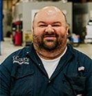 Man with a beard in a blue work shirt, smiling in a garage. | Emge's Car Craft Auto Services