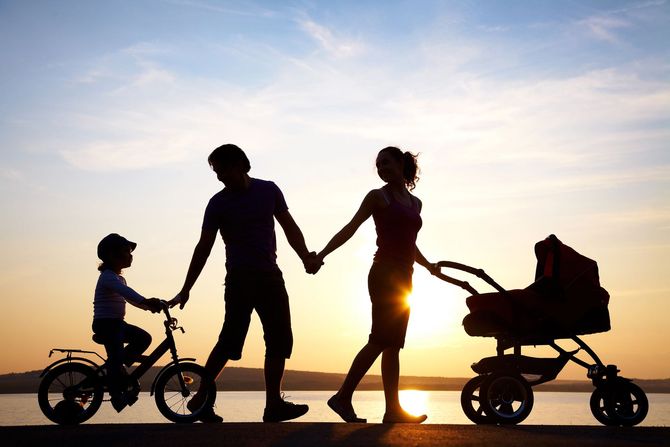 Una famiglia in silhouette che si tiene per mano con una bicicletta e un passeggino al tramonto su un sentiero sulla spiaggia.