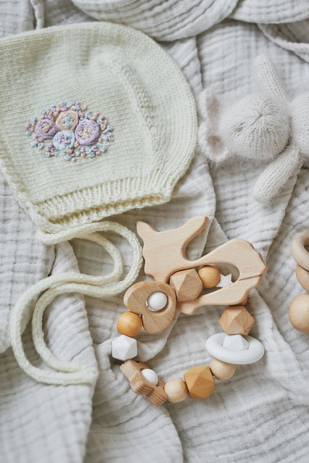 Cappellino da neonato lavorato a maglia, massaggiagengive in legno, clip per ciuccio e morbida copertina su uno sfondo di tessuto neutro.
