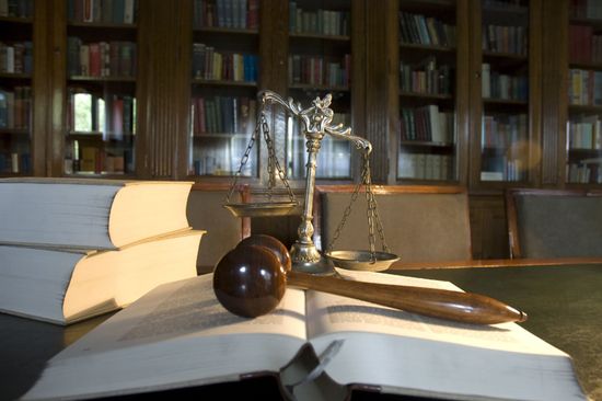 una bilancia a due piatti che pende dal lato destro, un libro aperto e sopra un martello di legno e sulla sinistra altri due grandi libri