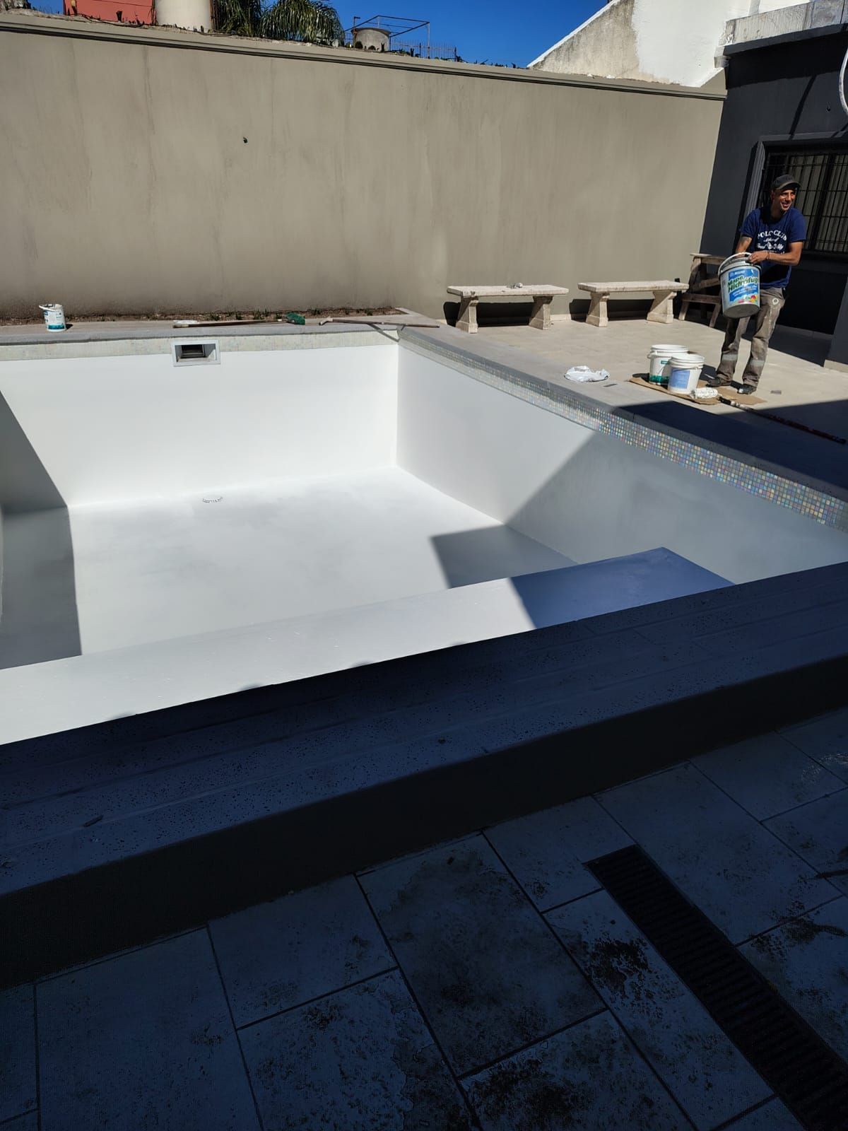 Piscina blanca vacía con un hombre de pie cerca. Paredes y azulejos grises rodean la piscina bajo un cielo azul.