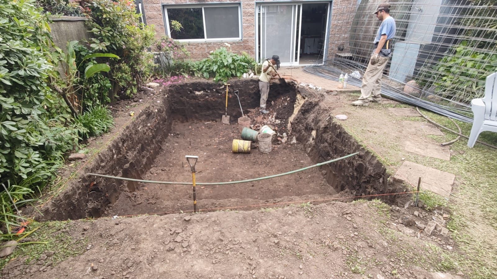 Personas excavando un hoyo rectangular en un patio trasero para un estanque; se observan herramientas y cubos.