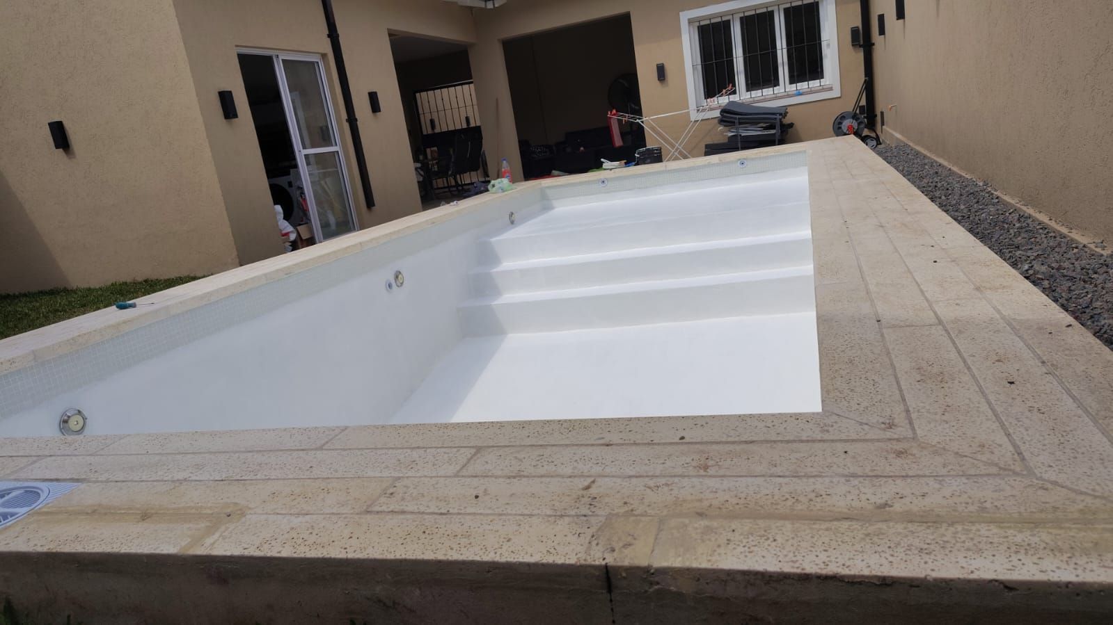 Piscina rectangular, blanca y vacía, con escalones, rodeada de pavimento de color claro y una casa al fondo.