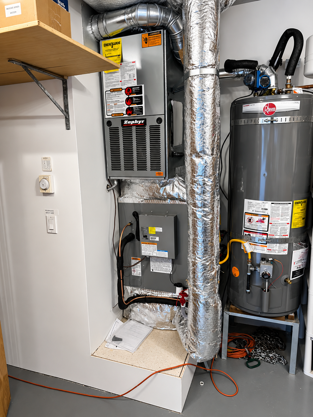 Utility closet with furnace, water heater, and exposed insulated ductwork next to a shelf and wall outlets