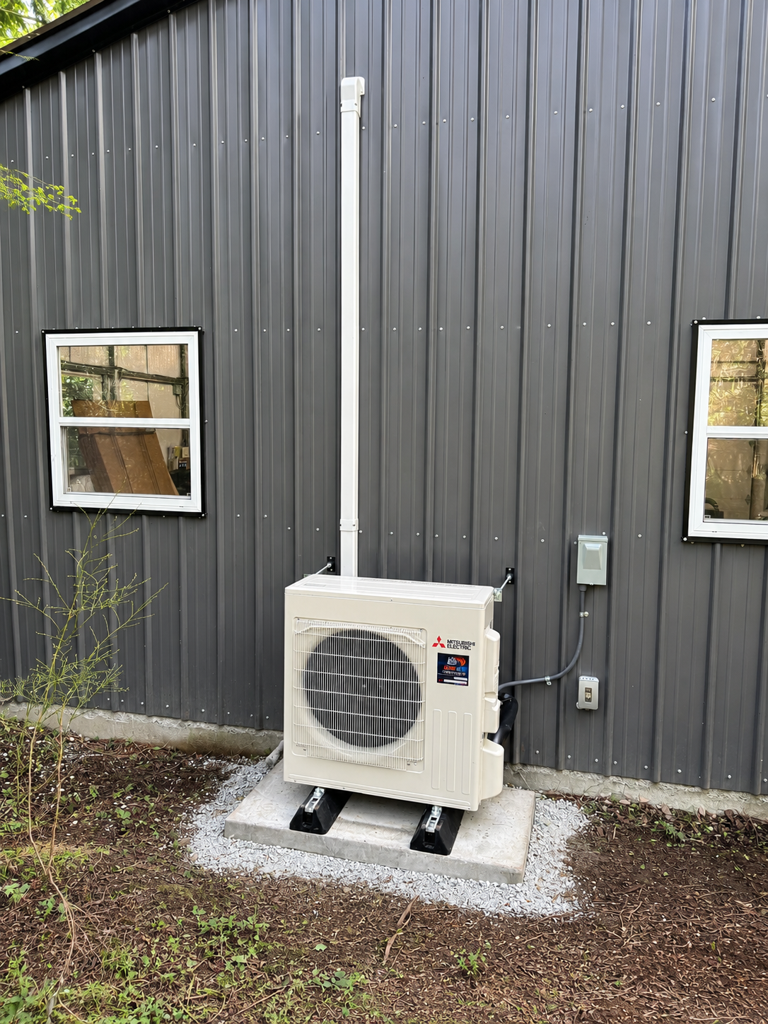 Outdoor heat pump unit against a dark gray building wall, with two windows and a conduit pipe.