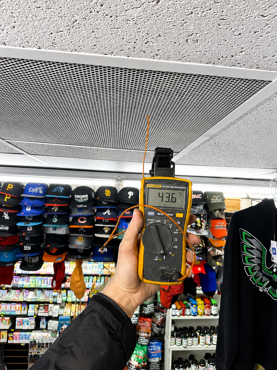 Hand holding a yellow handheld meter showing 43.6 in a store aisle with hats and clothing racks