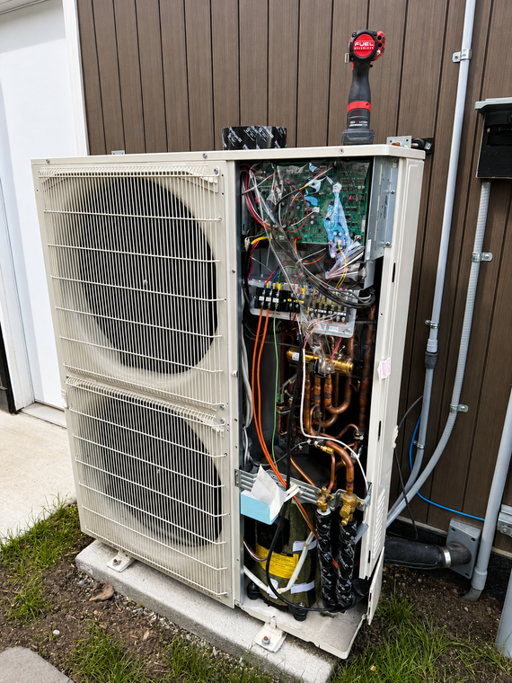 Outdoor HVAC unit with open panel, exposed wiring, and red tool on top beside a wooden fence