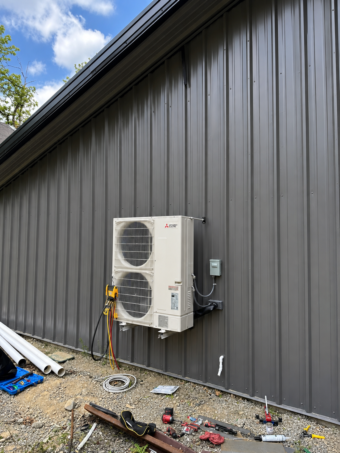 Outdoor heat pump mounted on a gray metal building wall with tools and gravel below