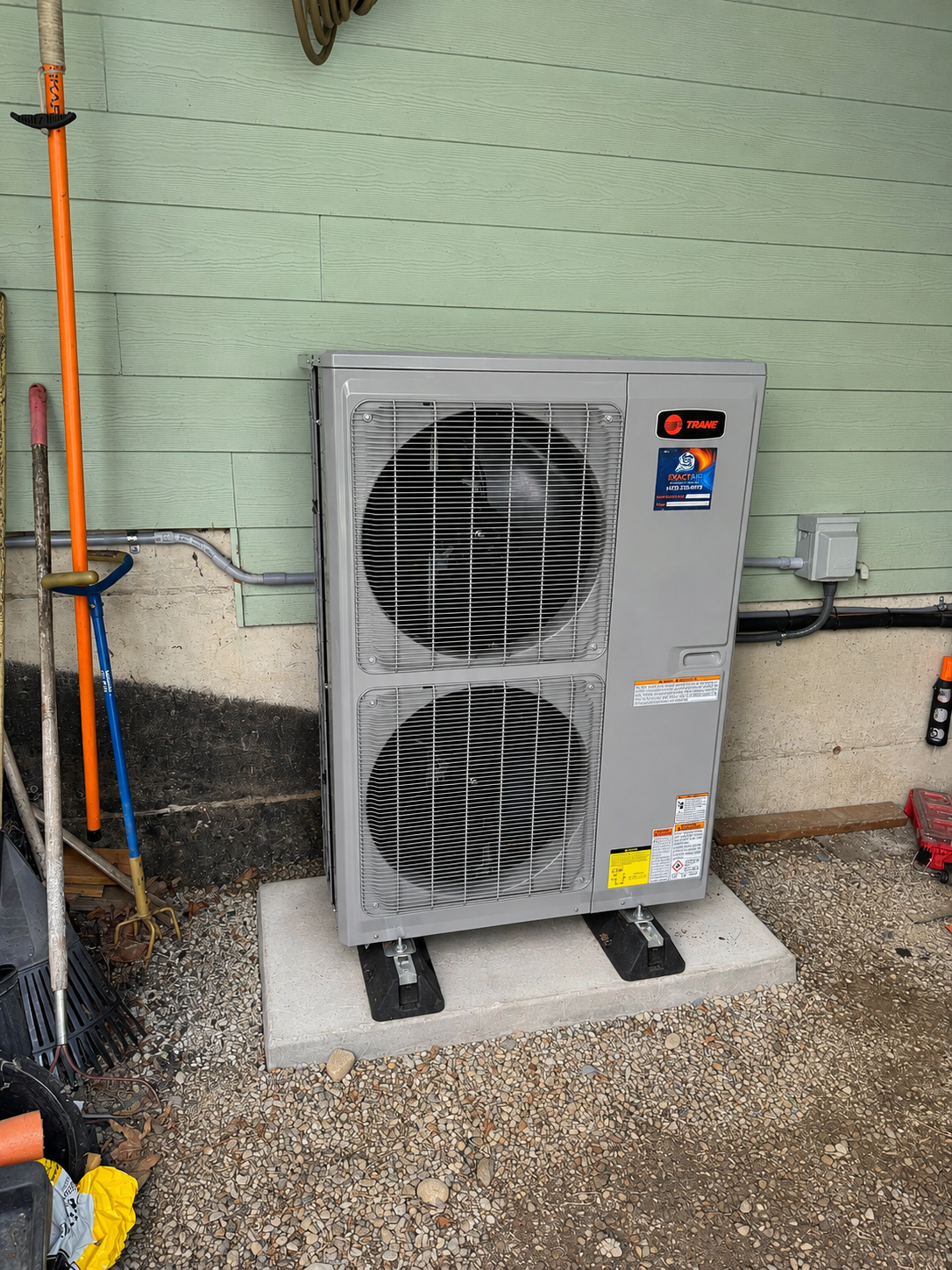 Outdoor HVAC unit beside a mint green wall, with orange and blue pipes and gravel ground.