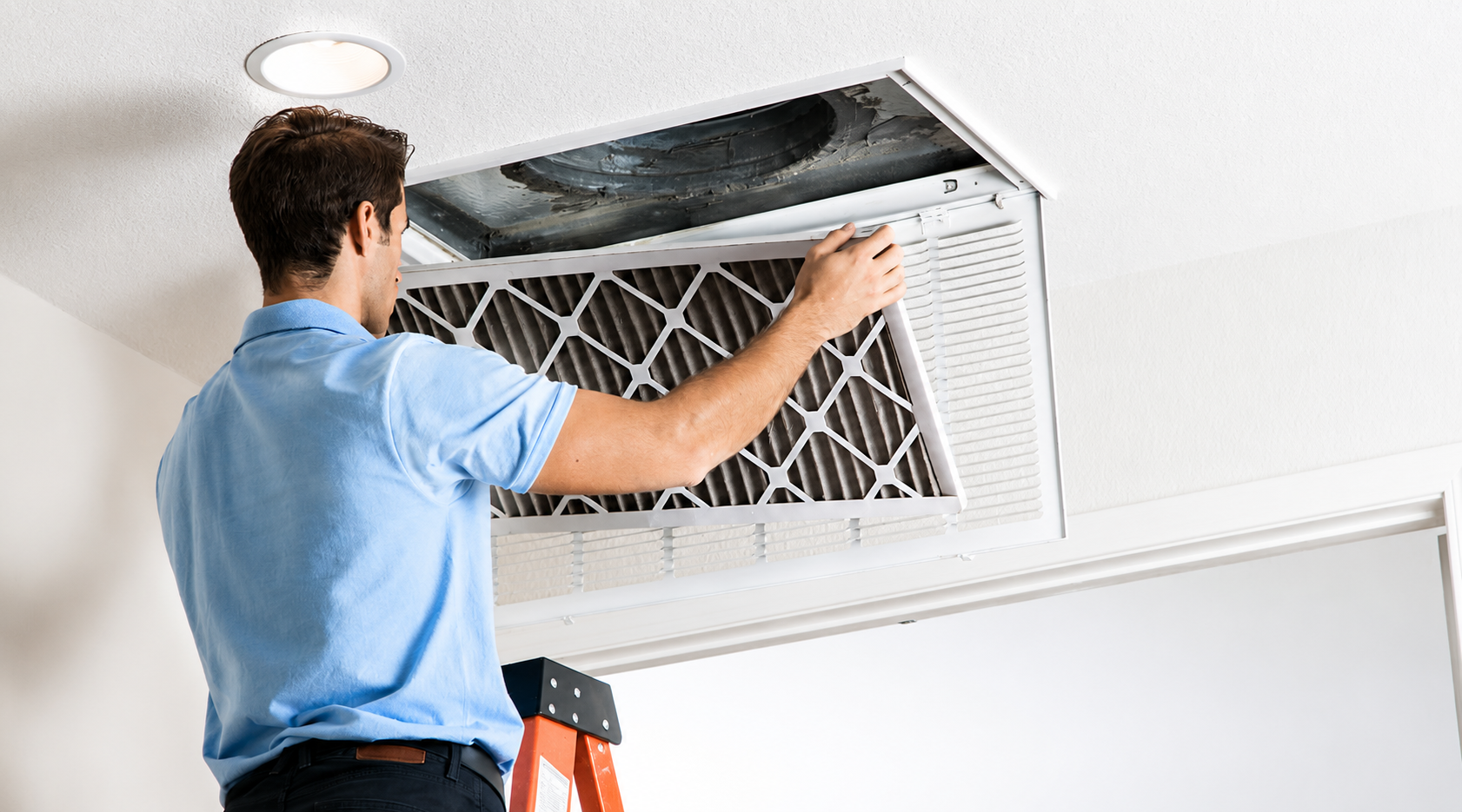 Technician cleaning a ceiling air vent in a white room while standing on a ladder