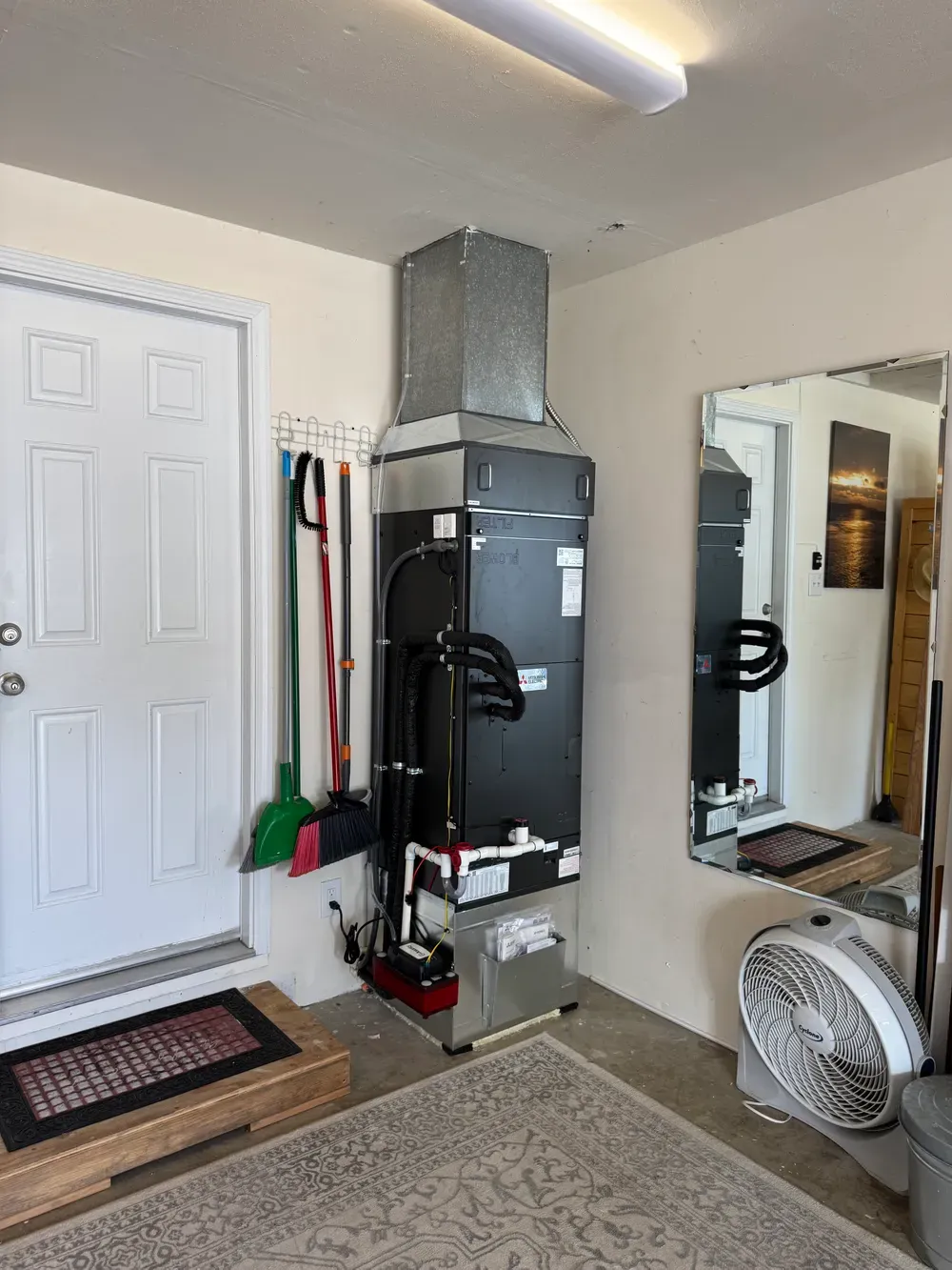 Small utility room with a wall-mounted black furnace, white door, mirror, fan, and doormat.