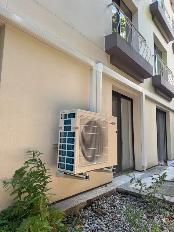 Outdoor wall-mounted air conditioner unit beside a beige building and balcony.