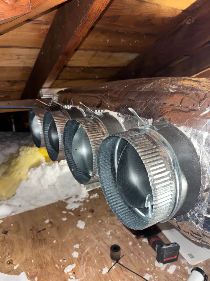 Insulated HVAC duct with four round metal vents running through an attic ceiling opening.