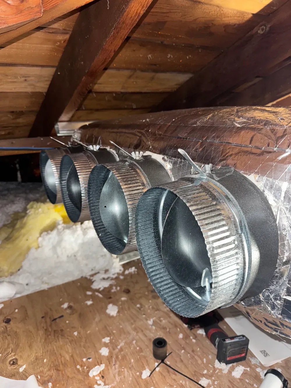 Flexible HVAC ducts in an attic, connected to a foil-wrapped main duct near wooden rafters.