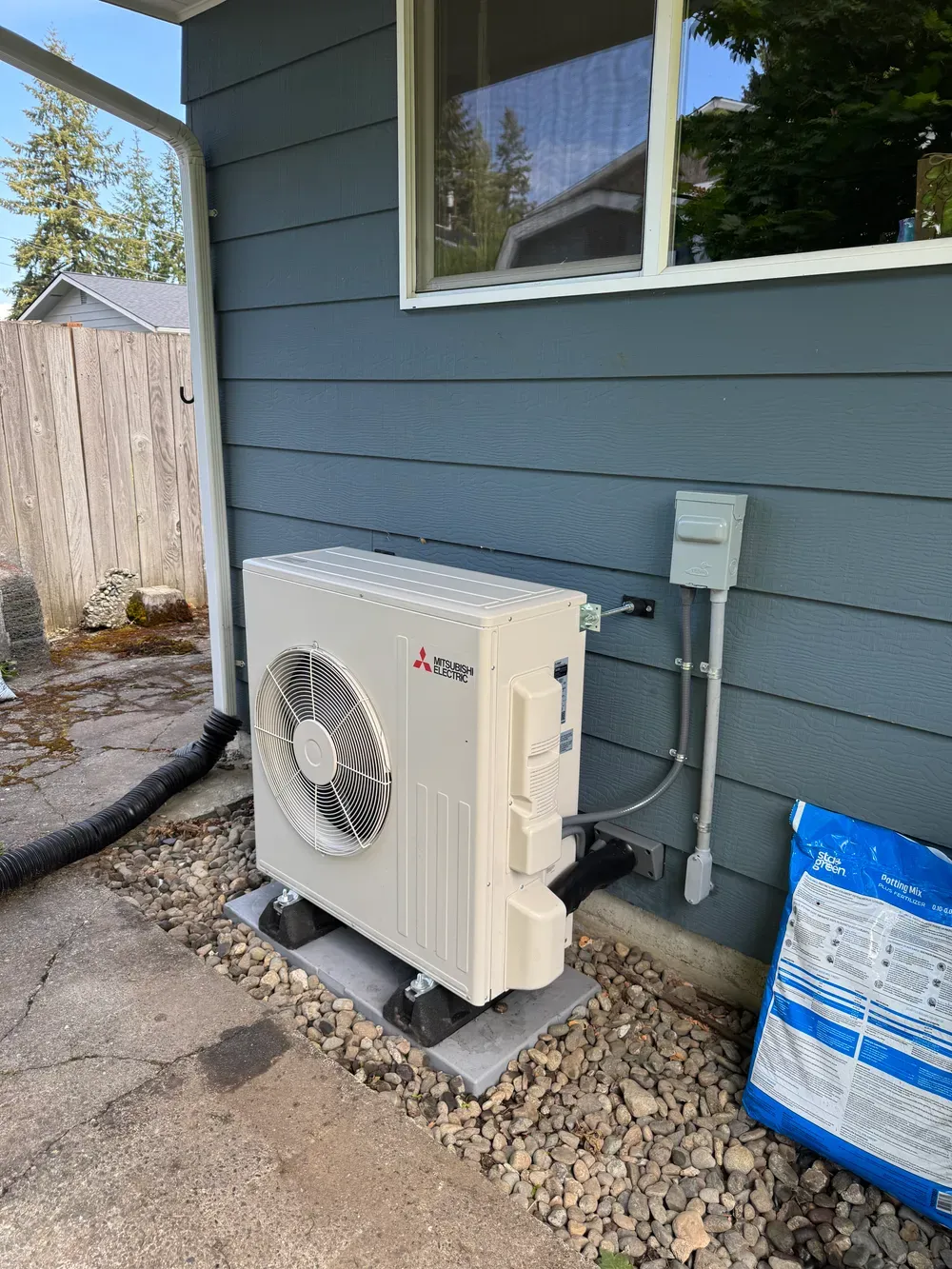 Outdoor heat pump unit mounted beside a gray house with conduit and gravel below a window