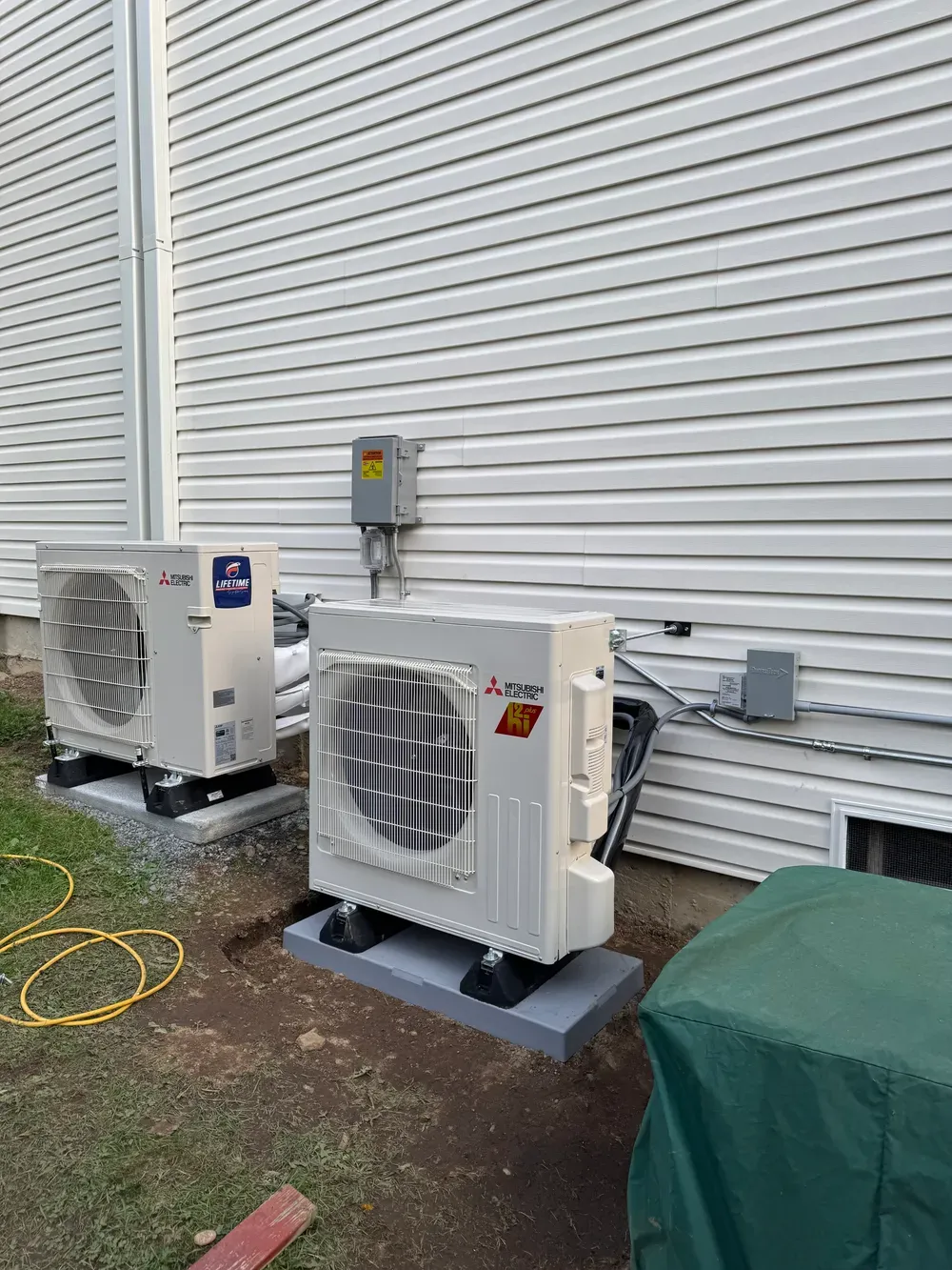 Two outdoor HVAC units beside a white building wall, with cords on the ground and a covered object nearby