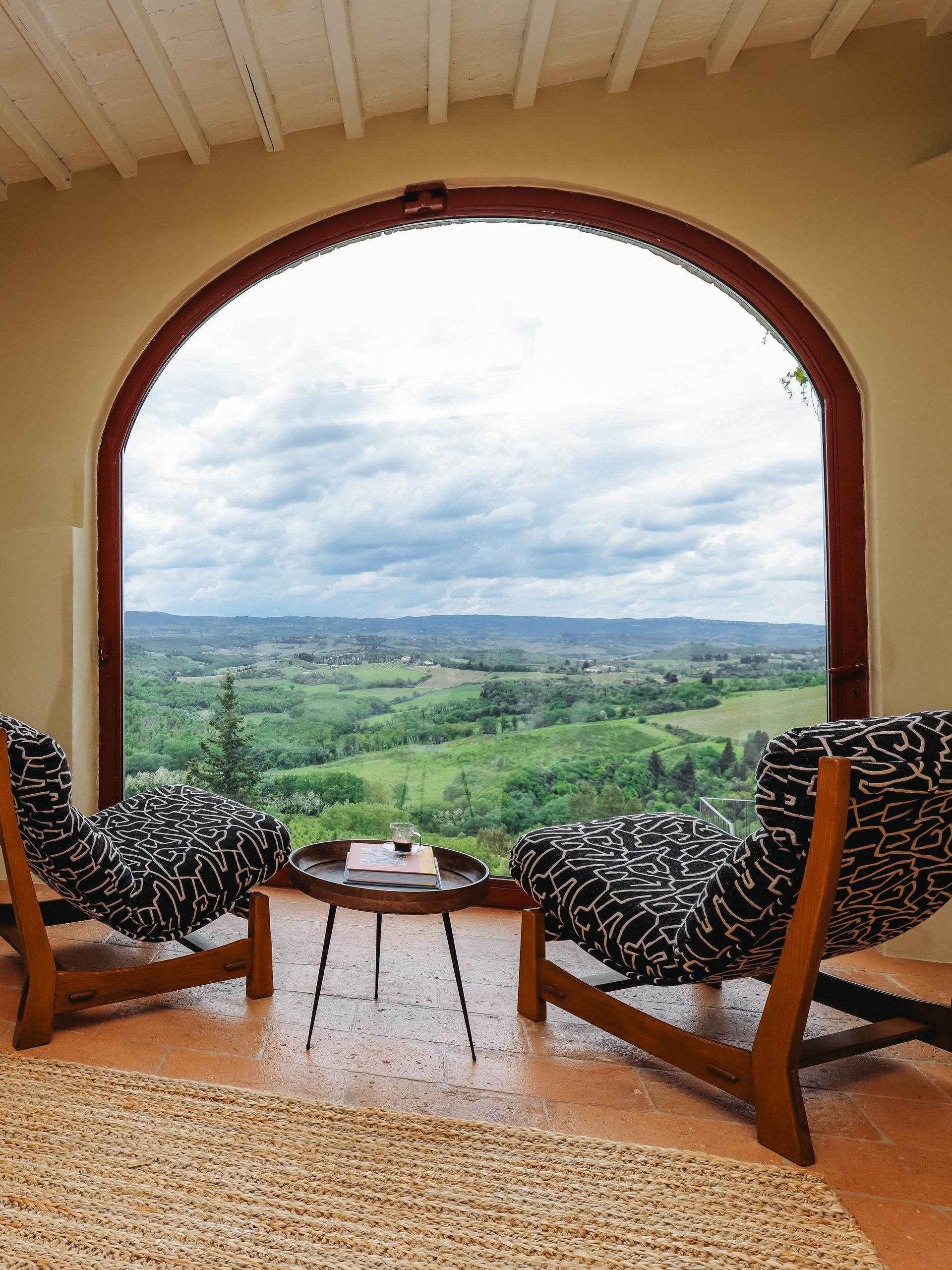 Two patterned chairs face a scenic landscape through an arched window.