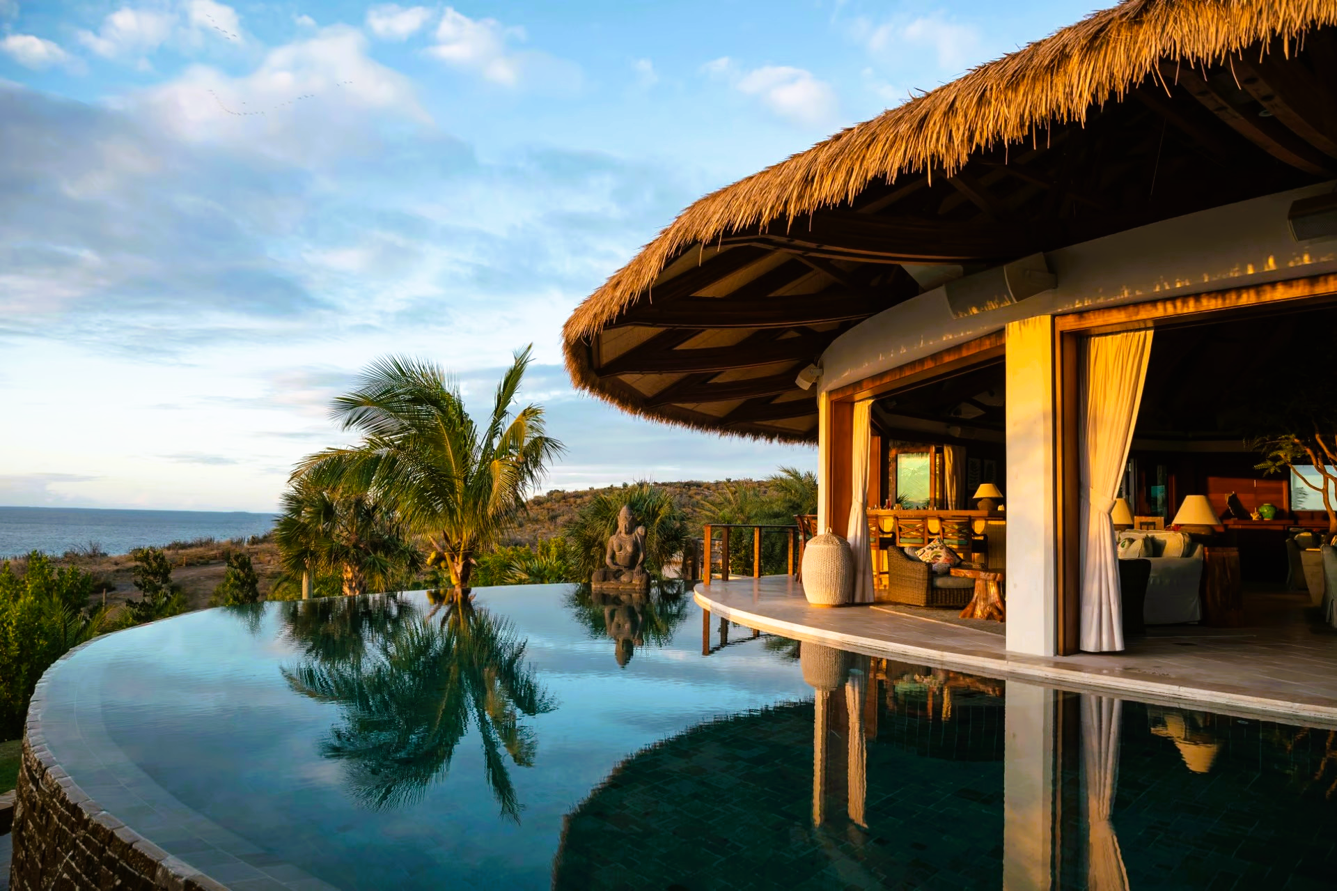 A house with a thatched roof has a swimming pool in front of it.