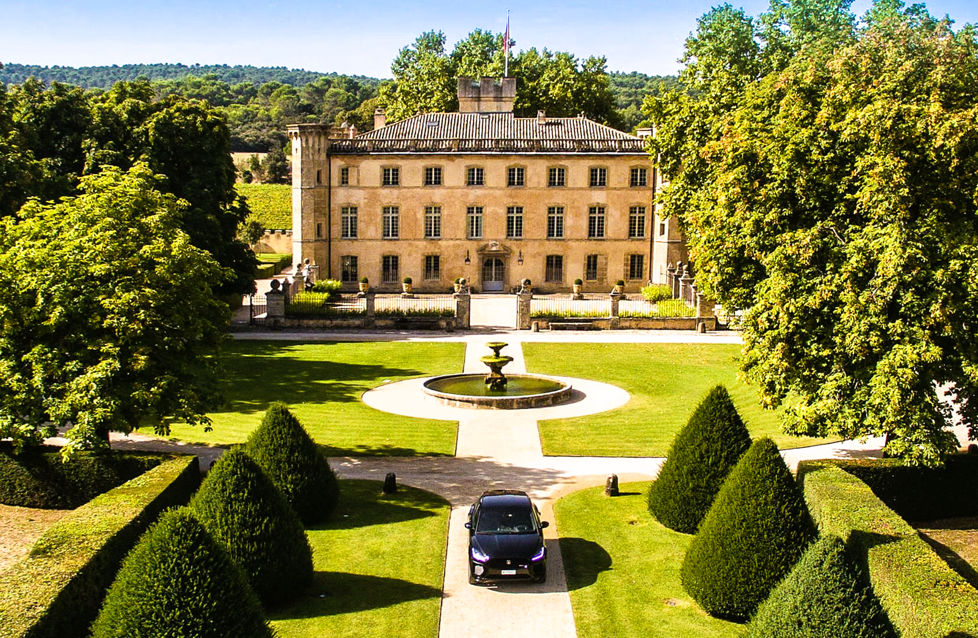 Black car parked in front of a grand stone chateau. Green trees and manicured lawn with fountain. Sunny day.