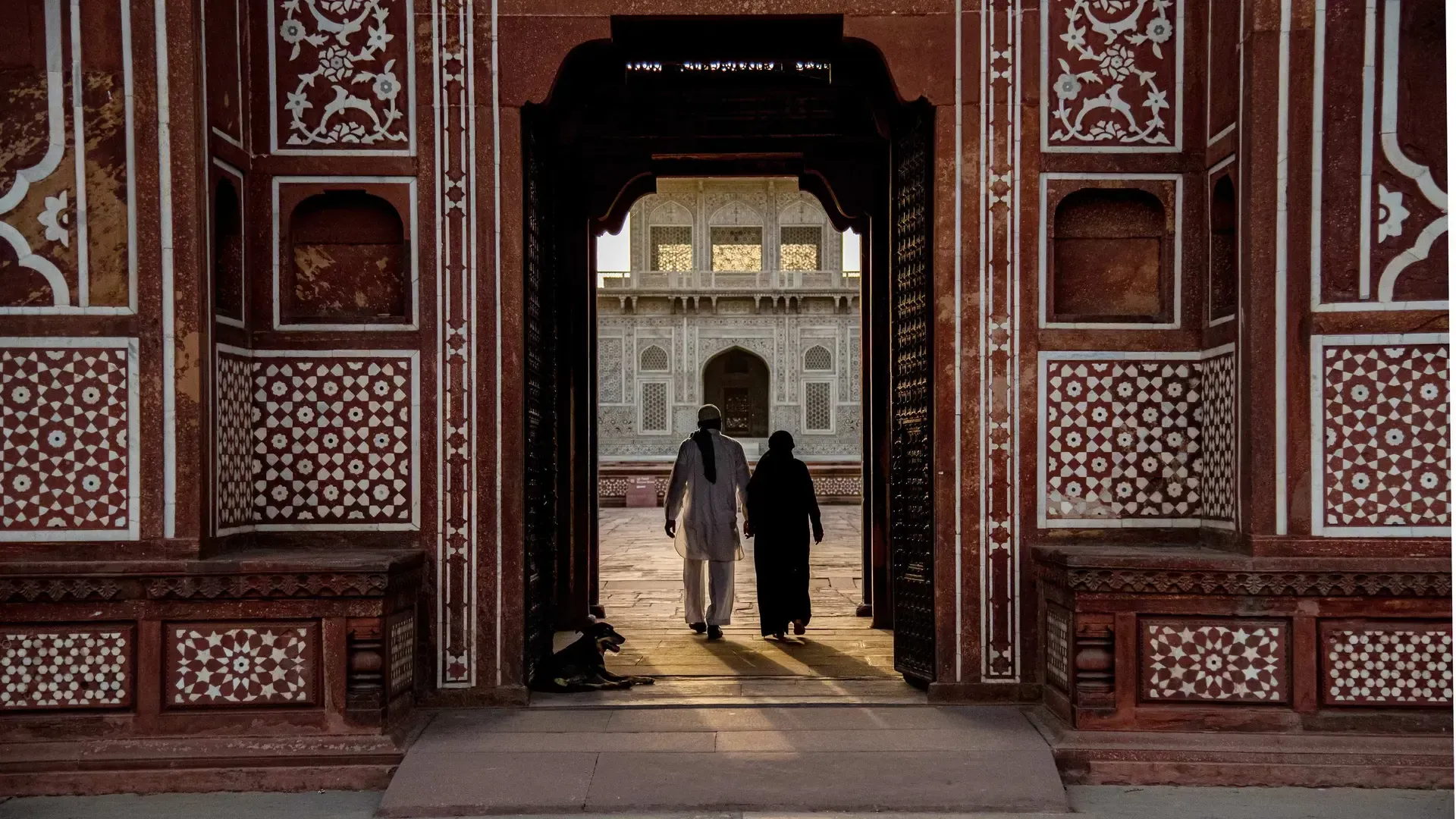Ornate archway frames two figures walking towards a bright, detailed building.