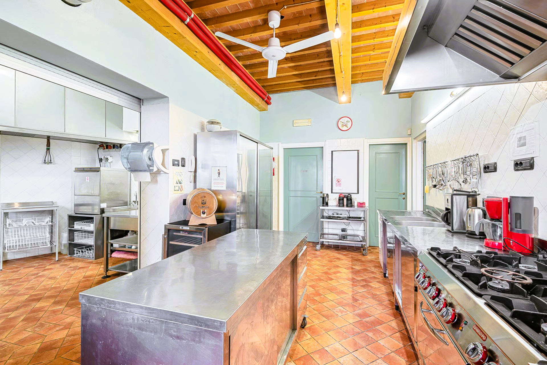 Commercial kitchen with stainless steel counters, large gas stove, brick floor, and wooden ceiling beams.