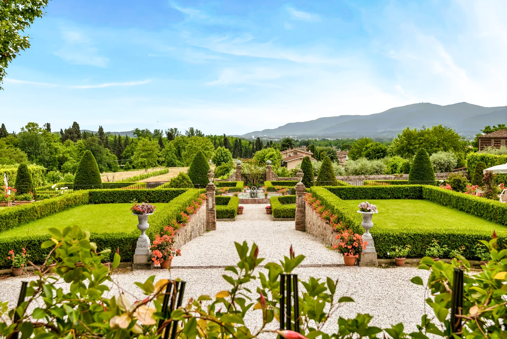 Formal garden with symmetrical hedges, stone paths, flower pots, and a mountain backdrop.