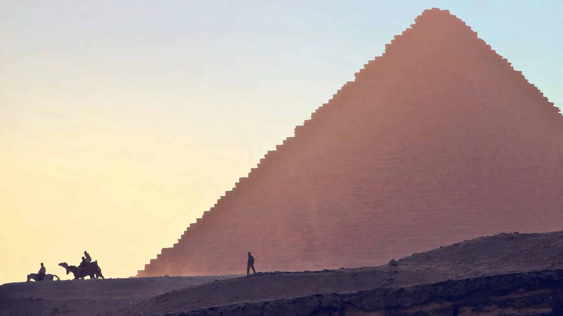 Pyramid silhouetted by sunset with people on camels and walking in the foreground.
