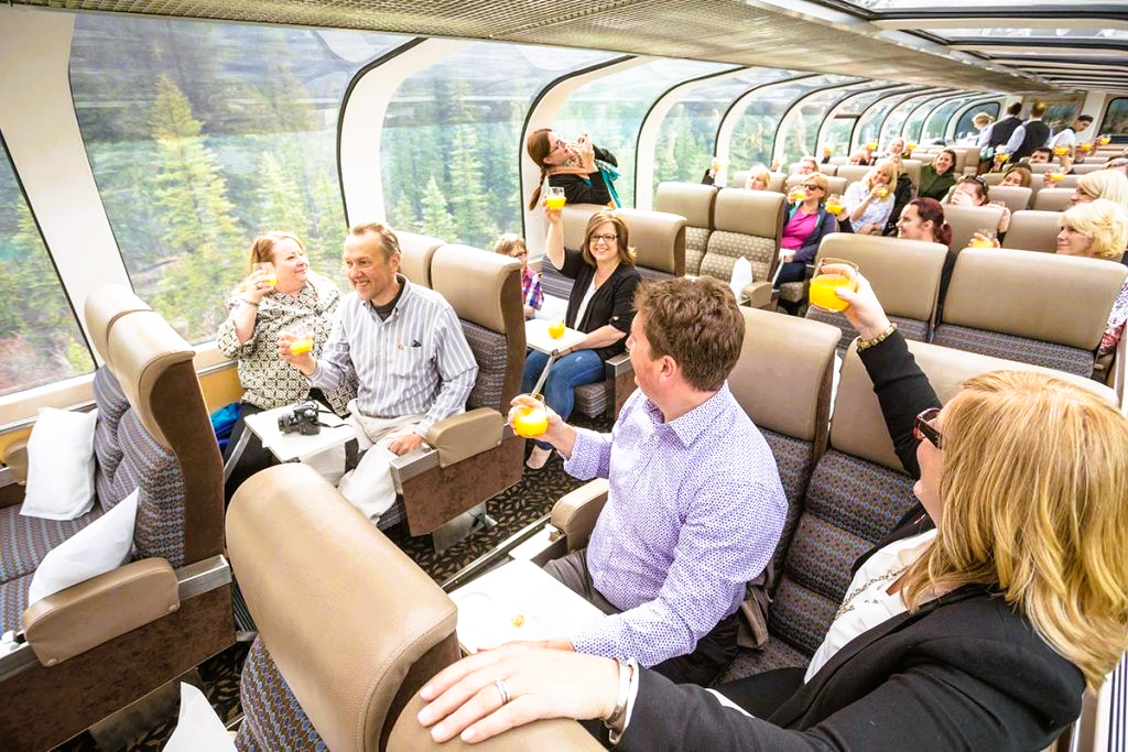 People on a luxury train are toasting with drinks, in a car with large windows.