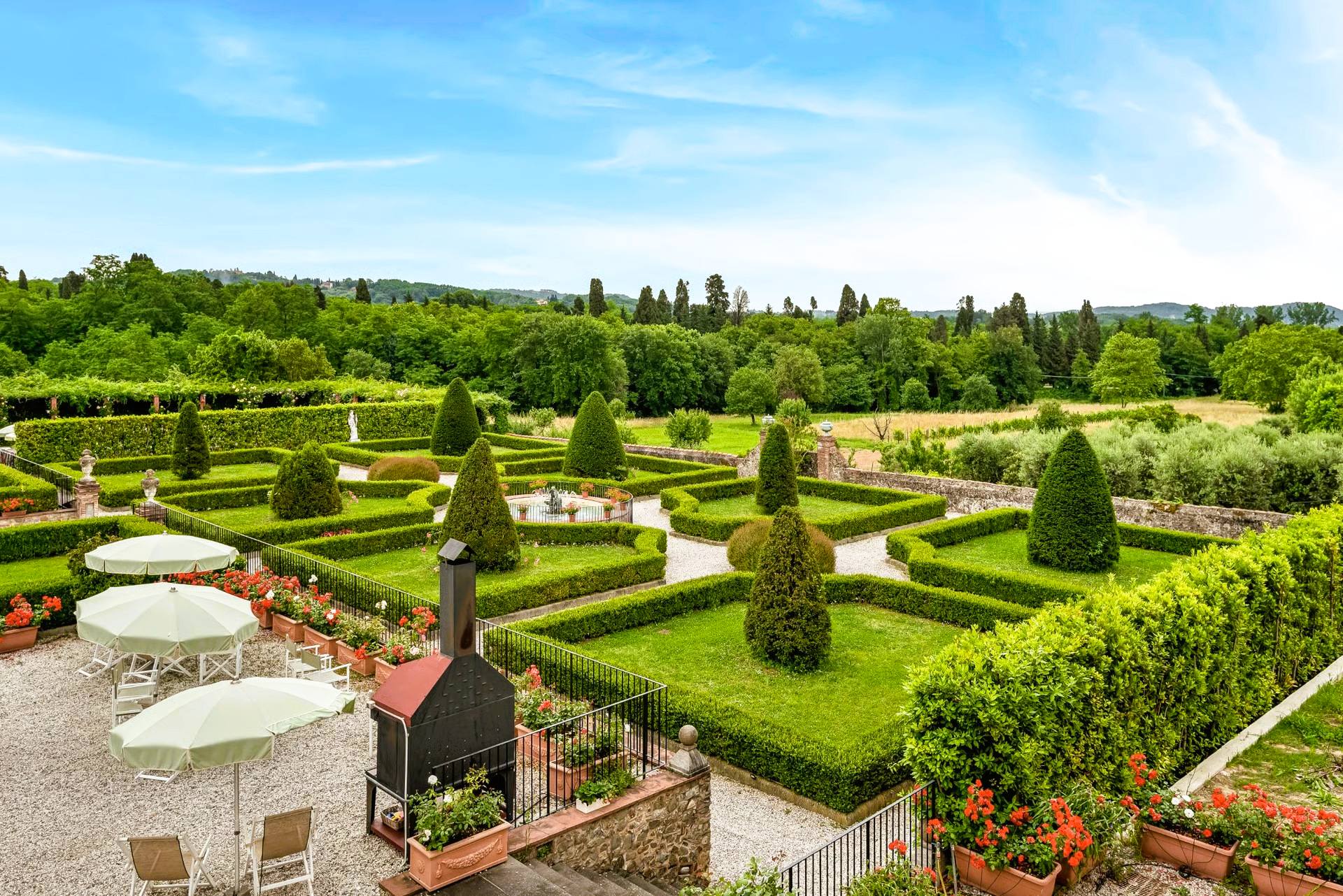 Formal garden with geometric hedges, sculptures, and trees under a blue sky.