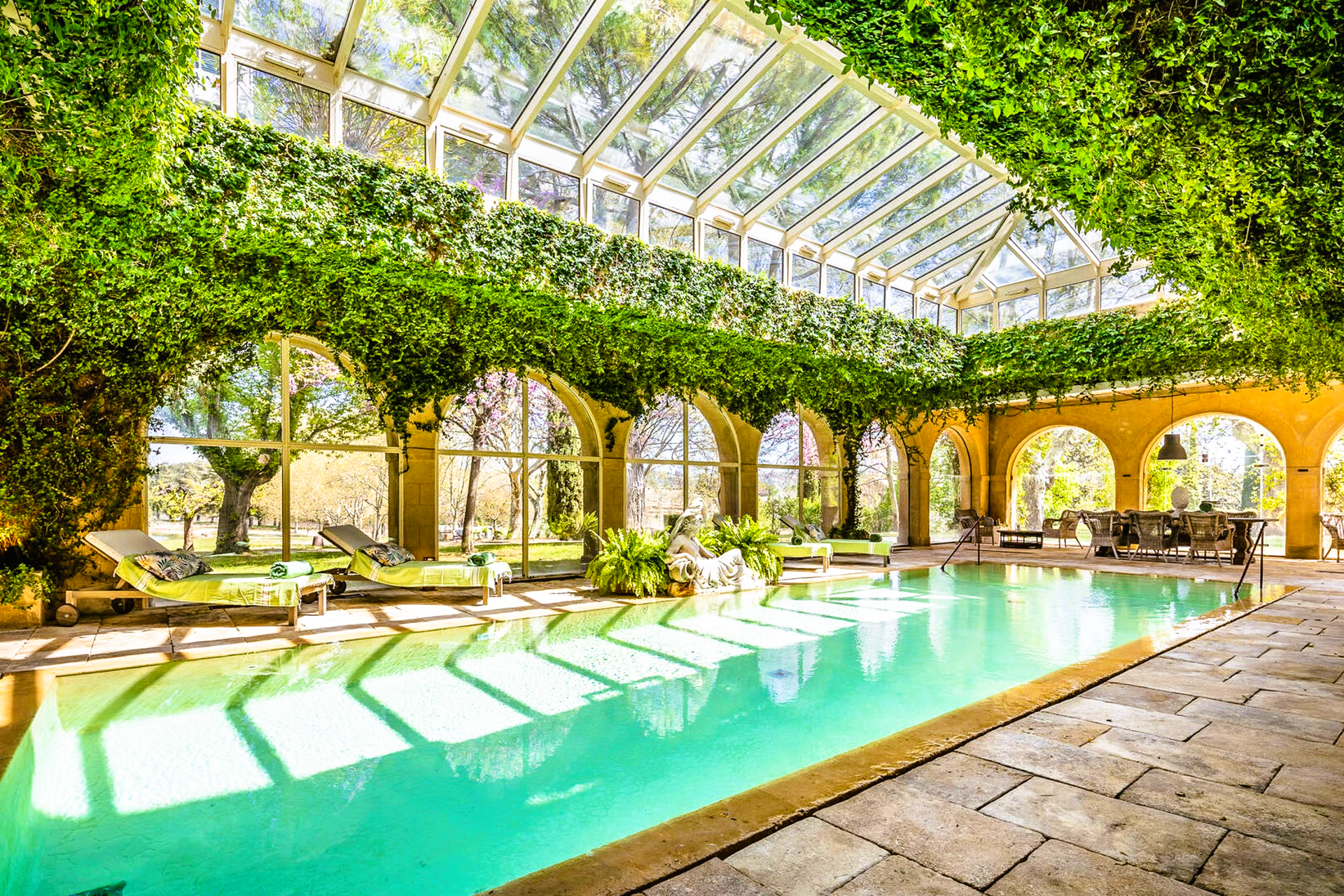 Indoor pool with arched windows, glass roof, and greenery. Sunlight reflects on the turquoise water.