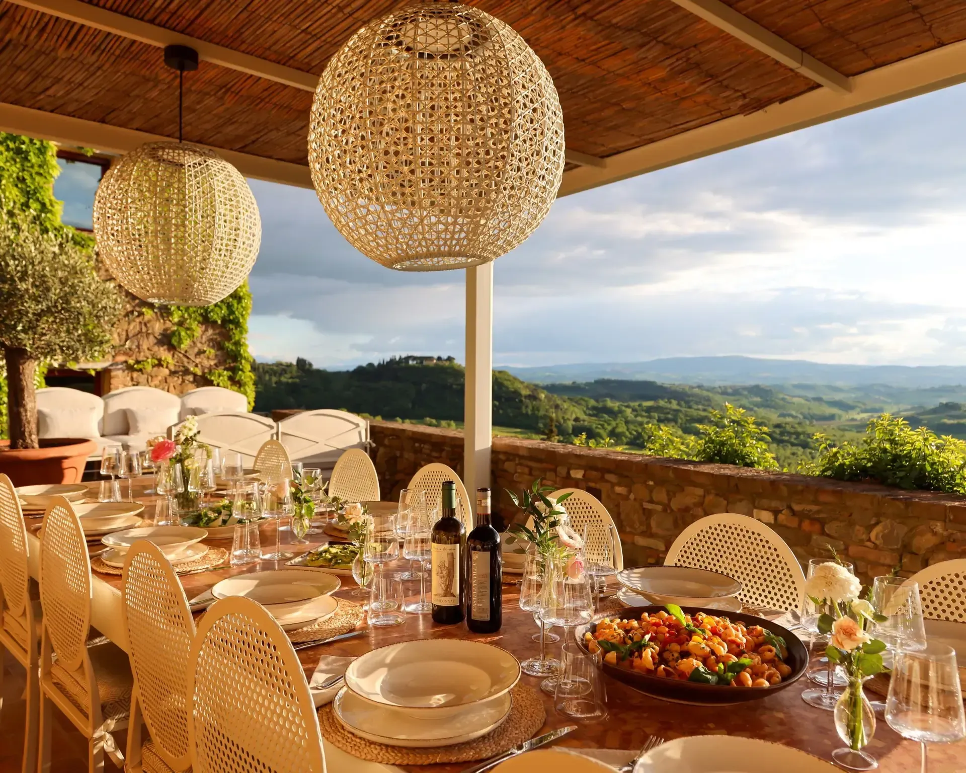Outdoor dining table set for a meal, with a scenic view. Two hanging lights and lush greenery.