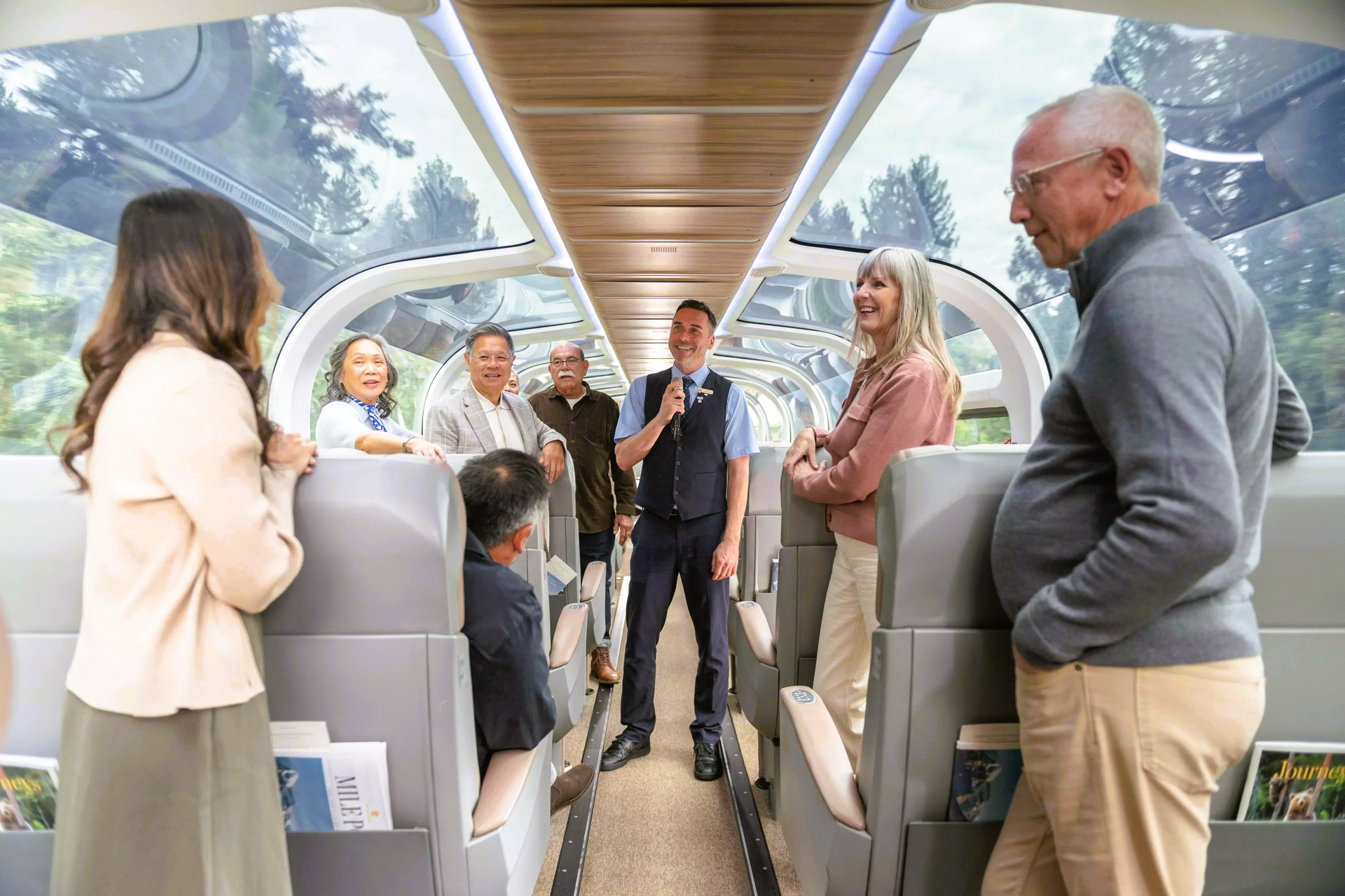 Inside a train car: Group of diverse seniors and conductor interacting, enjoying views through glass roof.