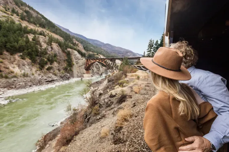 Couple on train, looking out at river and bridge. Brown and green landscape.