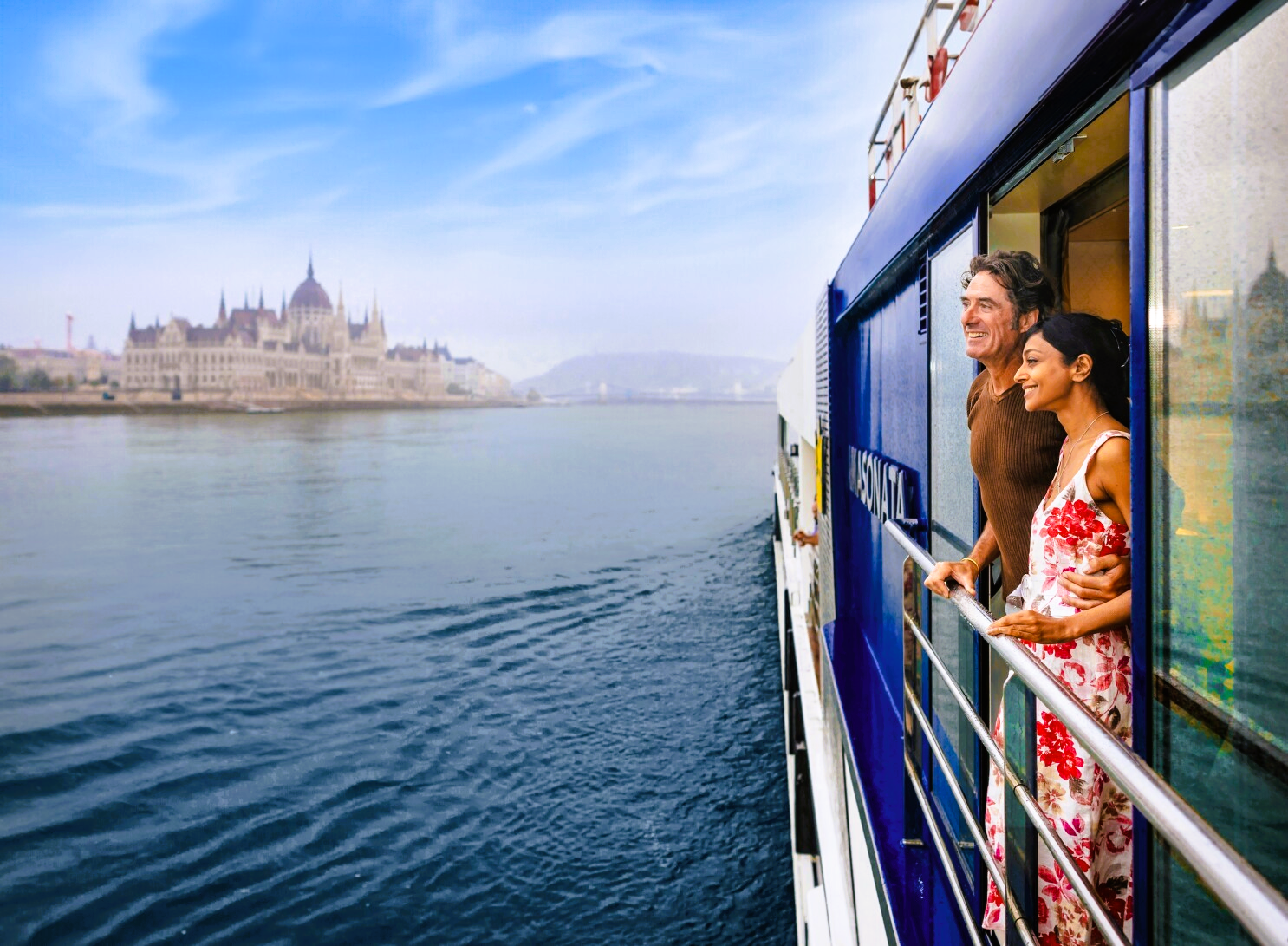 Couple on boat, looking at Budapest Parliament building along the Danube River on a sunny day.