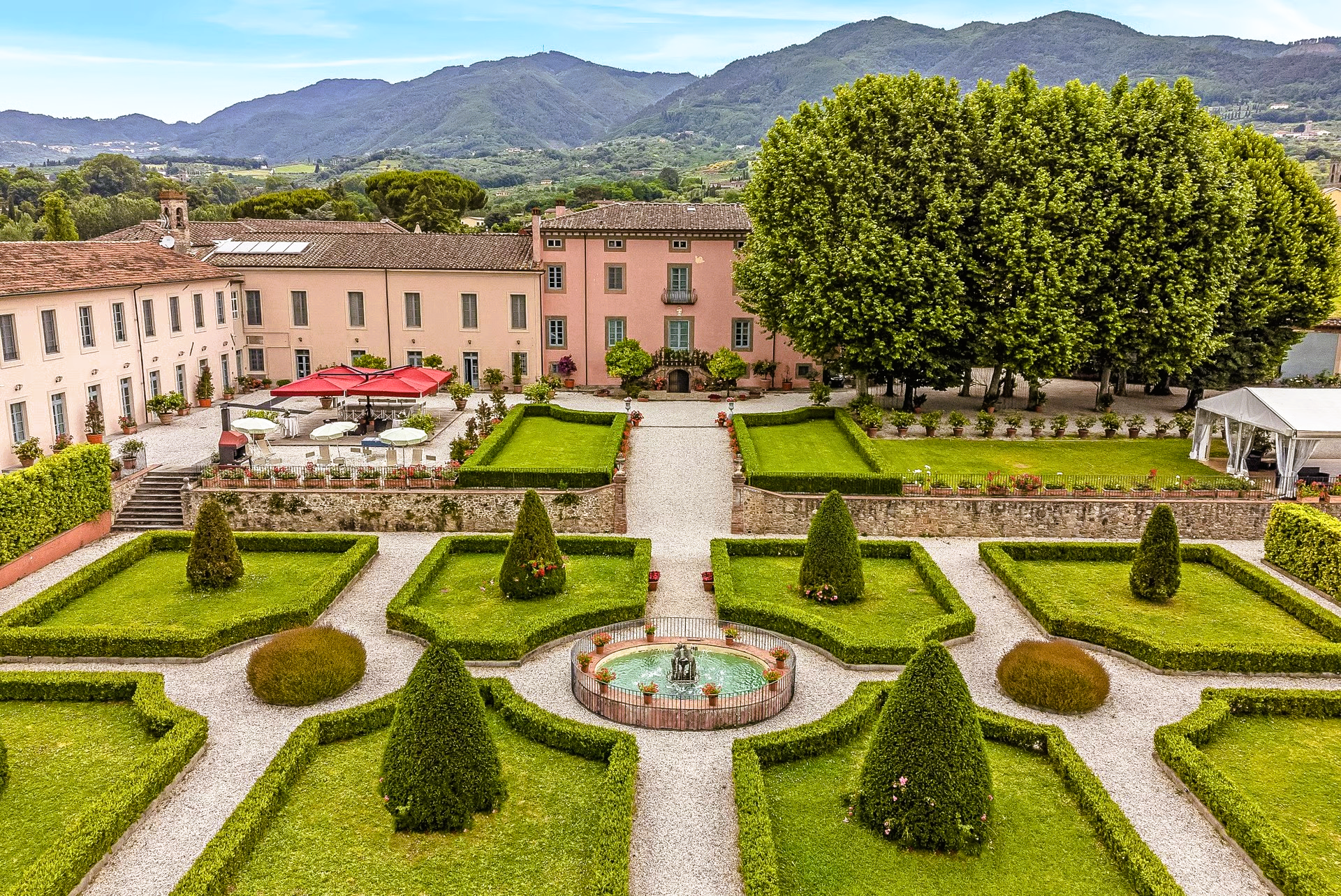 Formal garden with a fountain, geometric hedges, and a pink building in the background, mountains visible.