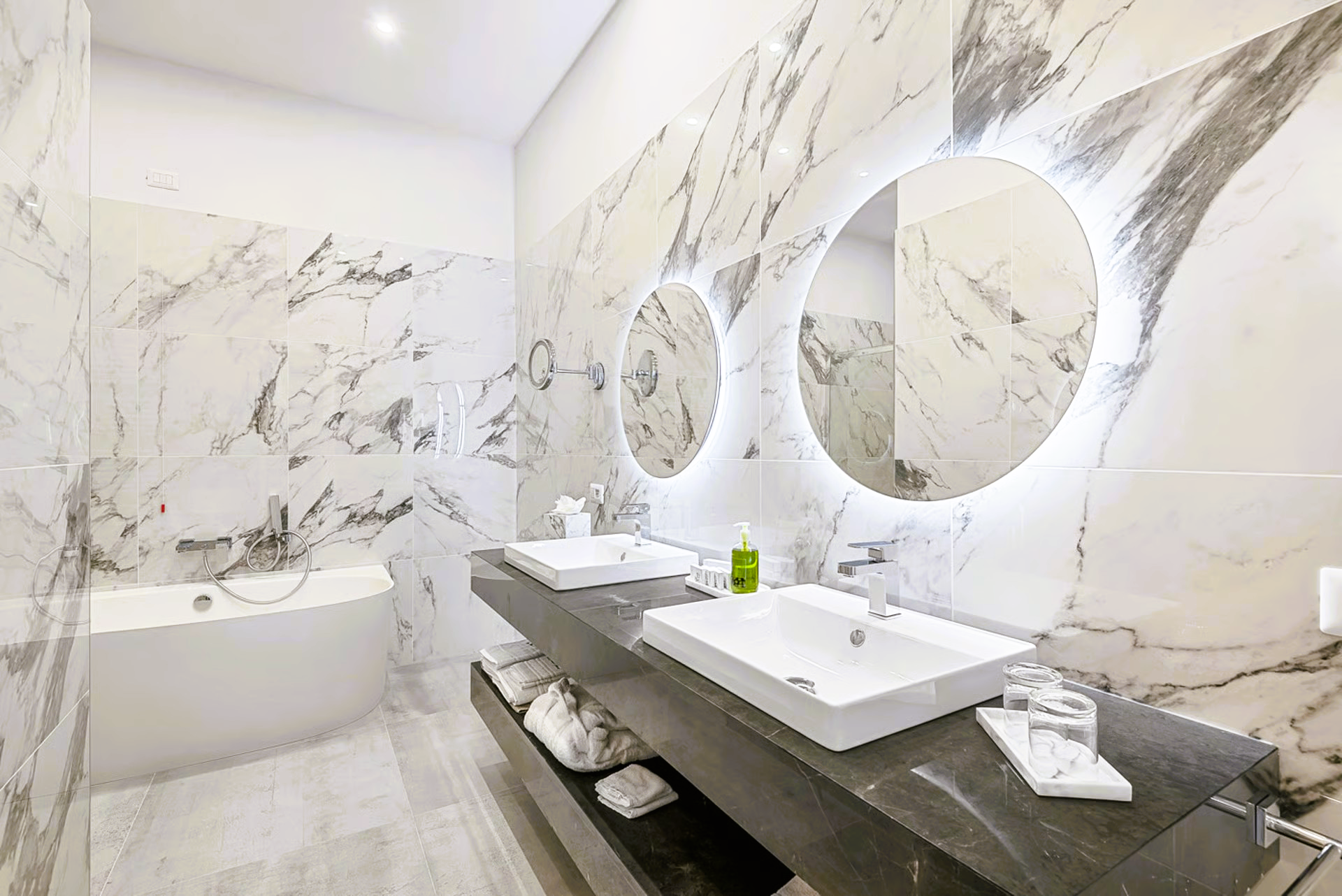 Modern bathroom with marble walls, two sinks, and illuminated mirrors.