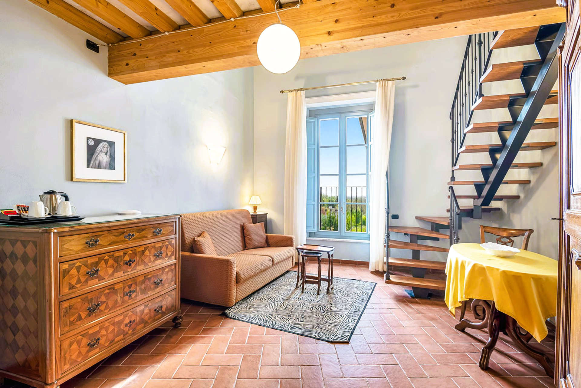 Cozy living area with a couch, dresser, and a staircase. Red tiled floors, white walls, wooden beams.