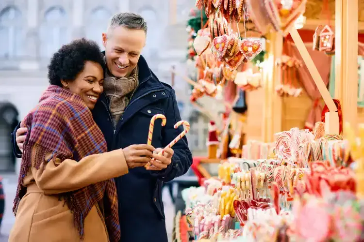 Couple at a Christmas market, smiling, holding candy canes. Colorful stalls, outdoor setting.