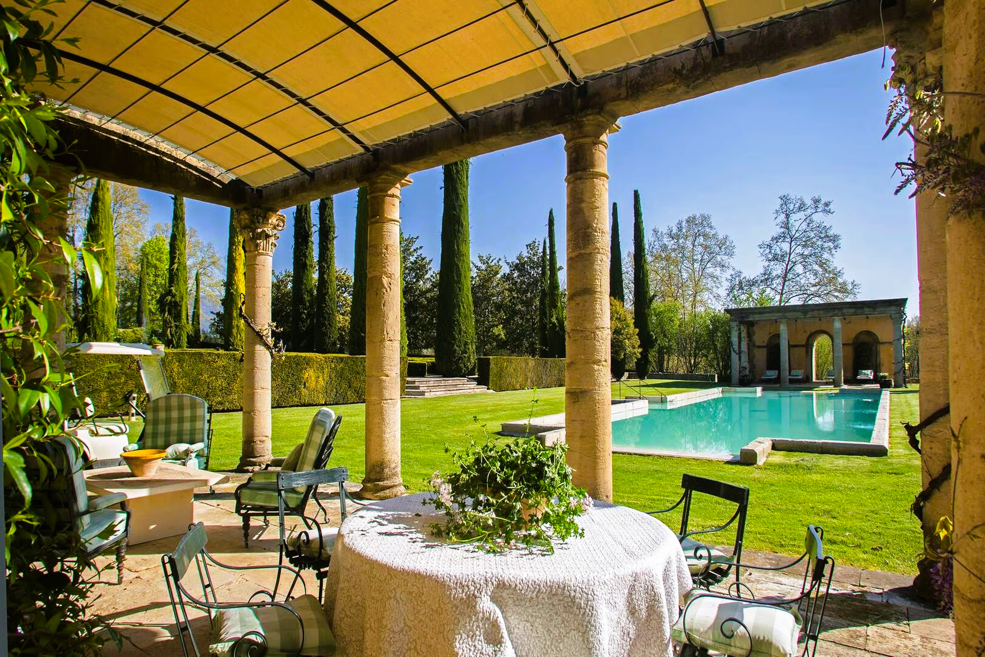 Patio overlooking pool and garden, with table, chairs, and tall trees against a bright sky.