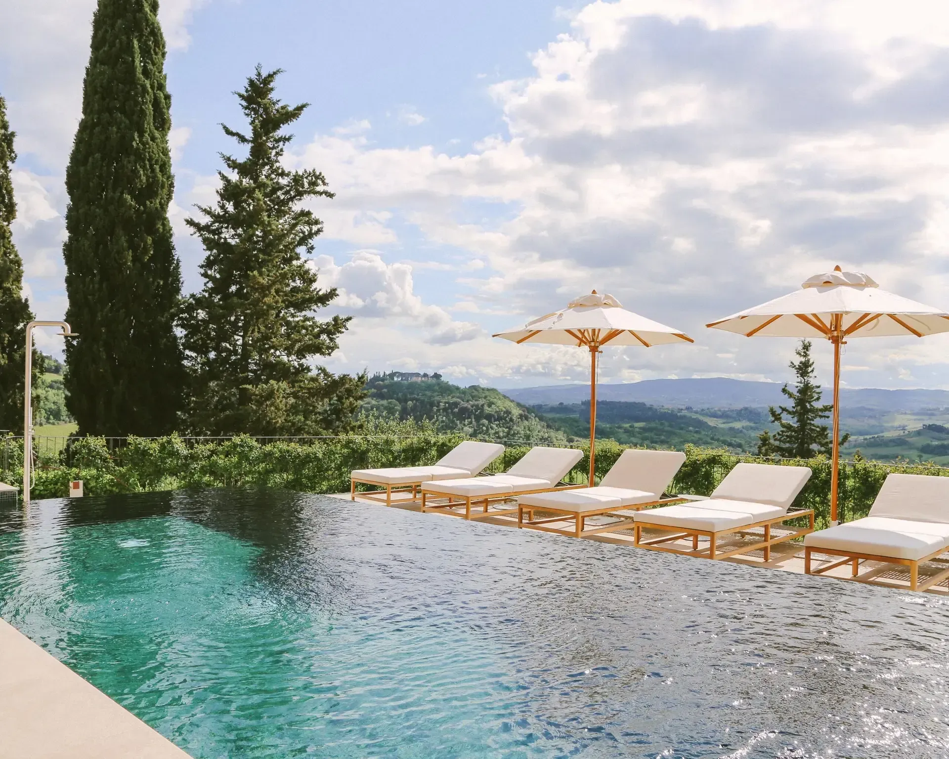 Infinity pool with lounge chairs and umbrellas overlooking rolling hills and vineyards under a cloudy sky.