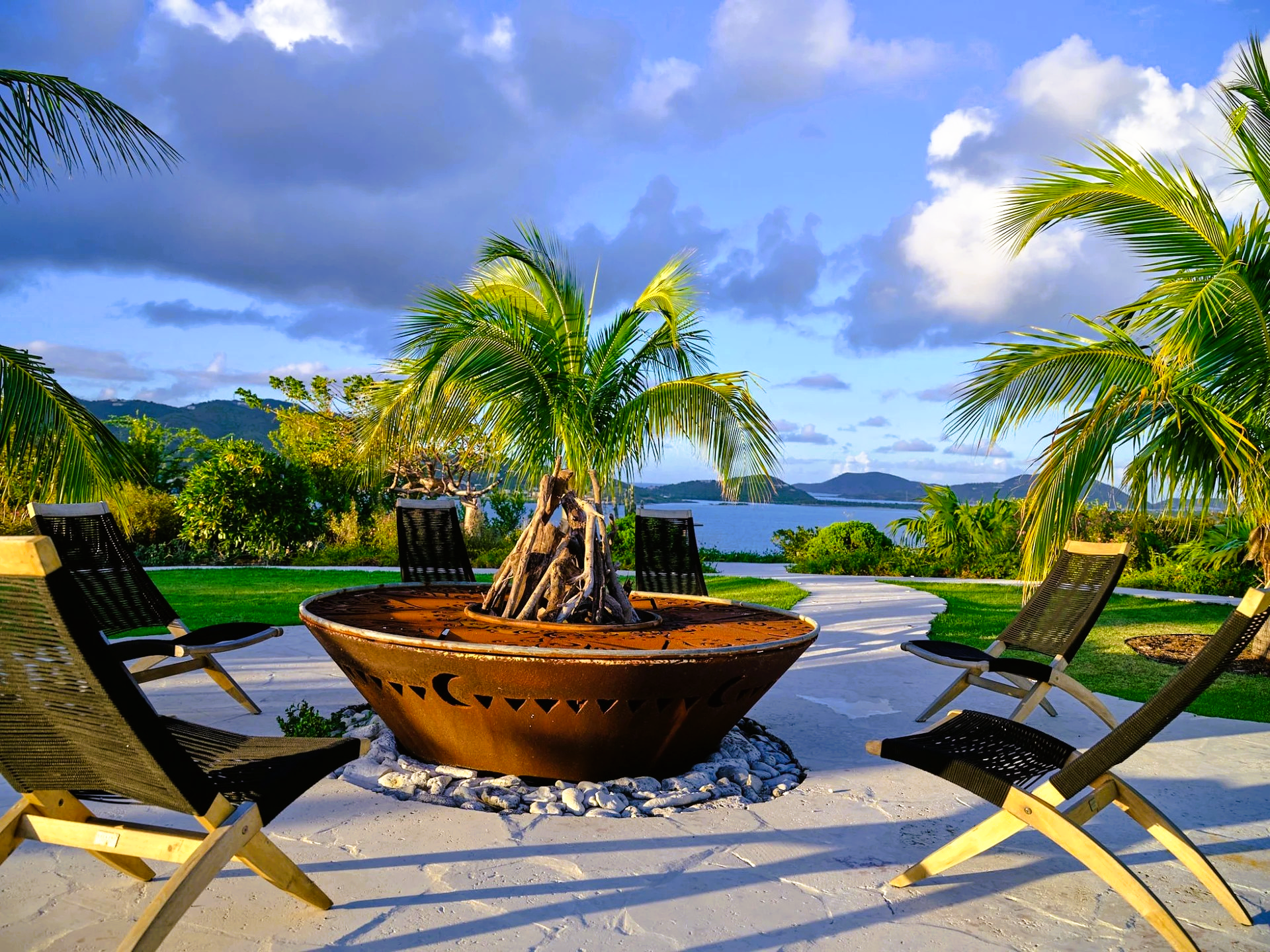 A fire pit is surrounded by chairs and palm trees on a patio.