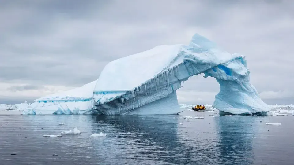 Iceberg with an arch formation over water under a cloudy sky. A small boat is near the arch.