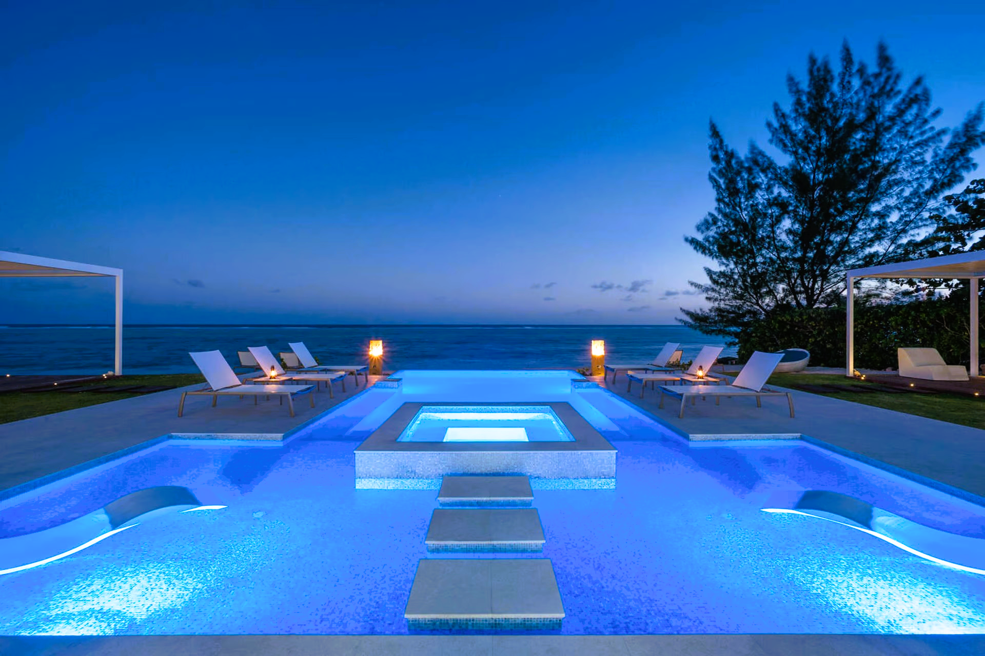 A large swimming pool is lit up at night with a view of the ocean.