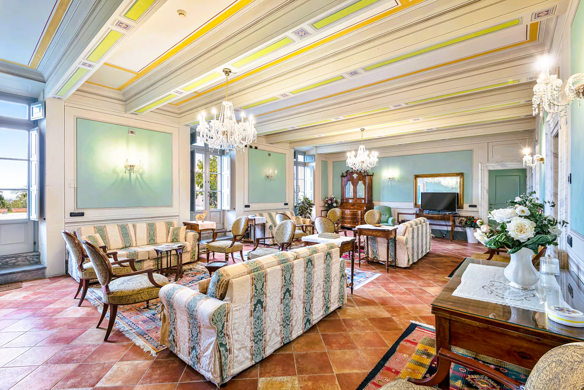 Spacious living room with couches, chandeliers, and antique furniture, terra cotta floor, blue walls, and light-colored ceiling.