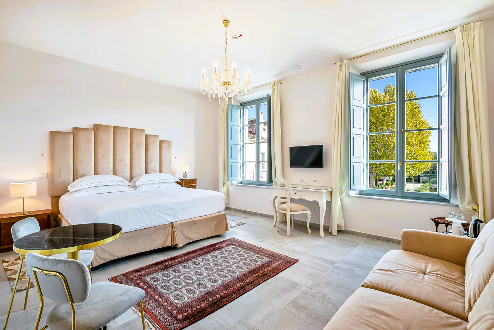 Elegant hotel room with a large bed, chandelier, windows, and seating area.