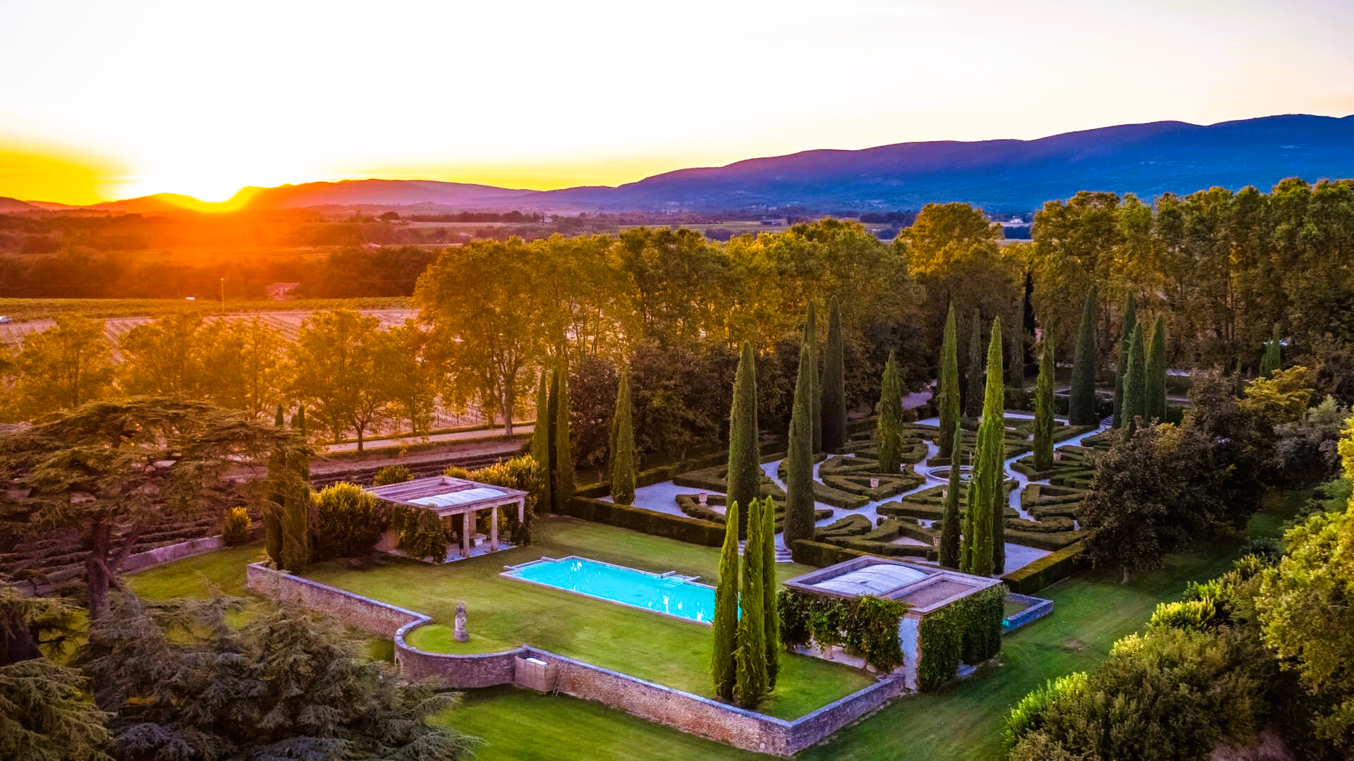 Sunset over a formal garden with pool, tall trees, and stone structures.